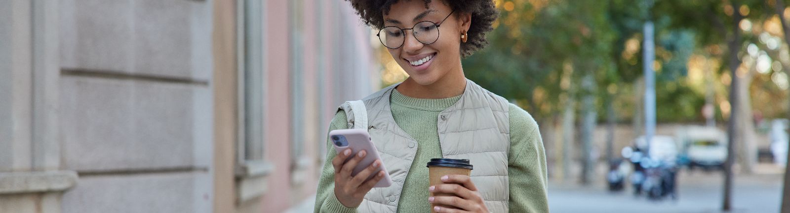 Smiling woman standing on a sidewalk holding a coffee cup and her phone